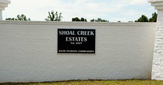 Black routed neighborhood sign for Shoal Creek Estates by David Pearson Communities, mounted on a white brick entrance wall.